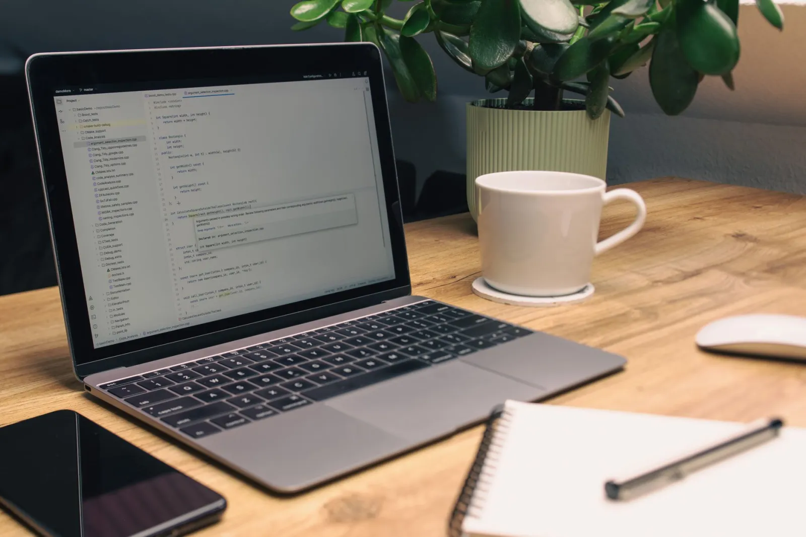 Laptop displaying code on a wooden desk with coffee, plant, and notebook.